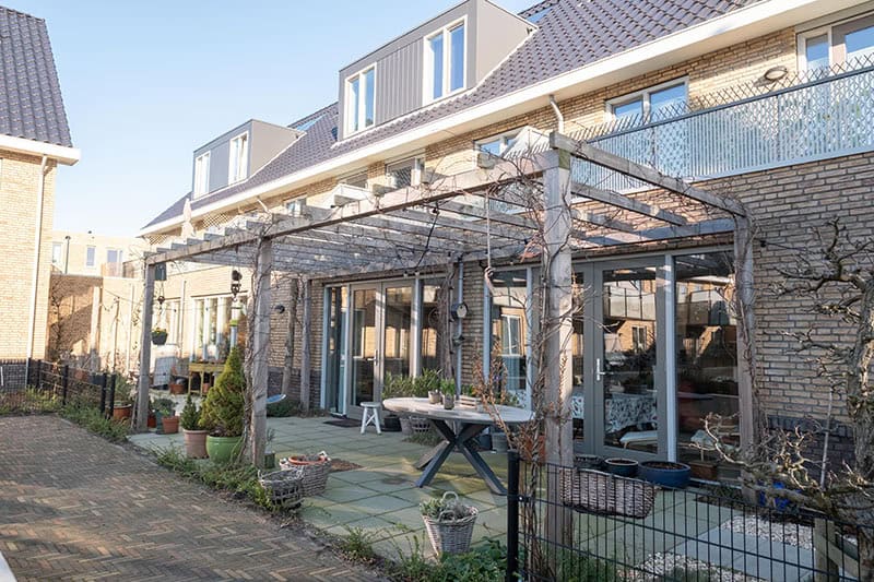 Achtertuin van een moderne rijtjeswoning met pergola, terras, plantenbakken en grote glazen deuren naar de woonkamer, op een zonnige dag.